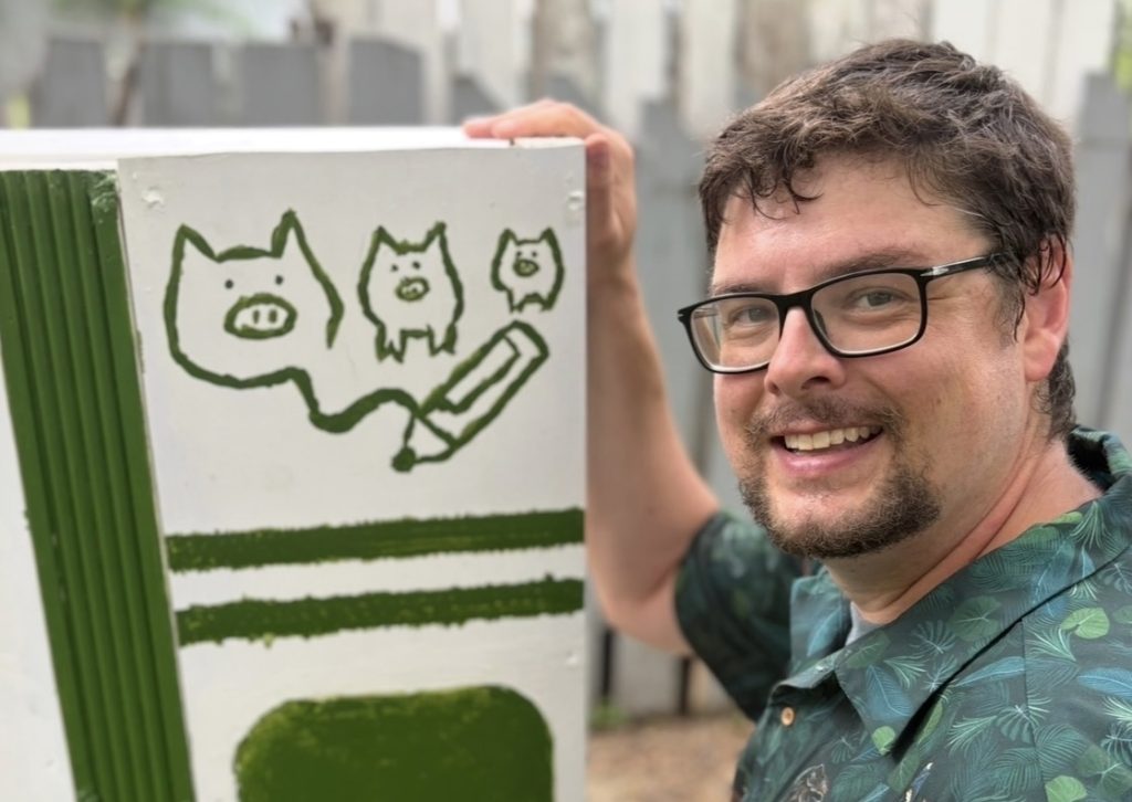 Kevin Kermack smiles next to a set piece for Storytelling Alive featuring the three little pigs and a pencil.